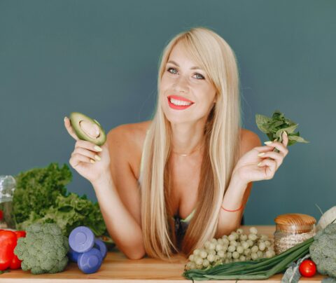 Blonde woman promotes healthy eating with fresh vegetables and fruits on display.